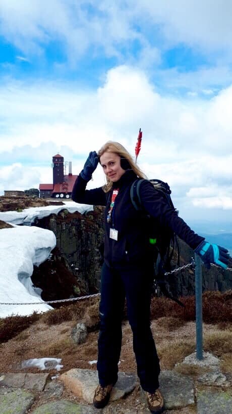 Kobieta w górach, ubrana w sportowy strój, z plecakiem, na tle budynku obserwatorium meteorologicznego na Śnieżce, z fragmentami śniegu i błękitnym niebem z chmurami.