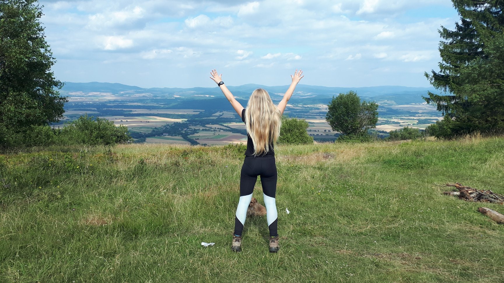 Kobieta z rozłożonymi rękami podziwia rozległy krajobraz w okolicach Oleśnicy, widok z góry na pola i wzgórza pod błękitnym niebem z chmurami.