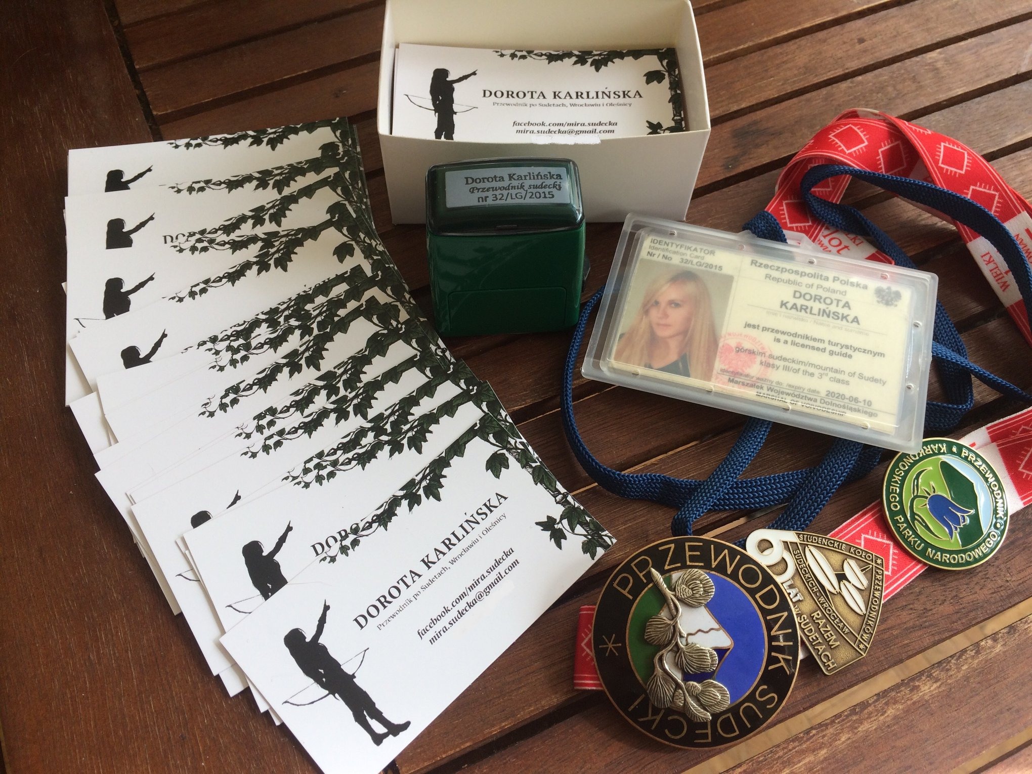 Wizytówki, pieczątka, identyfikator i odznaki przewodnika górskiego Doroty Karlińskiej, leżące na drewnianym stole, prezentujące jej kwalifikacje i dane kontaktowe.