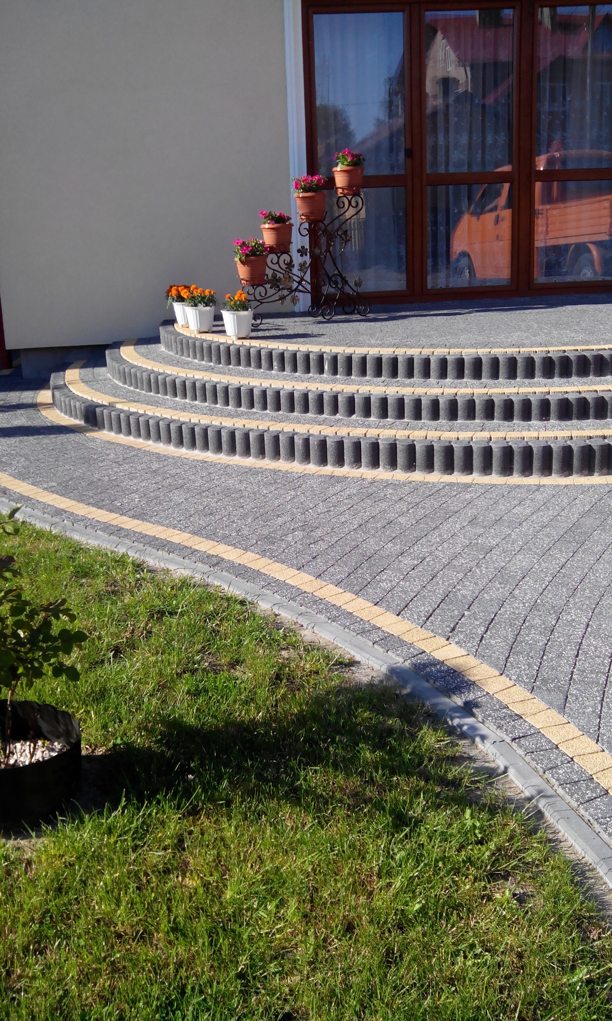 Kaskadowe schody wejściowe z kostki brukowej w odcieniach szarości, obrzeża w kolorze piaskowym, rabata kwiatowa z kwiatami w doniczkach na ozdobnym metalowym stojaku, trawnik z boku, w tle okno...