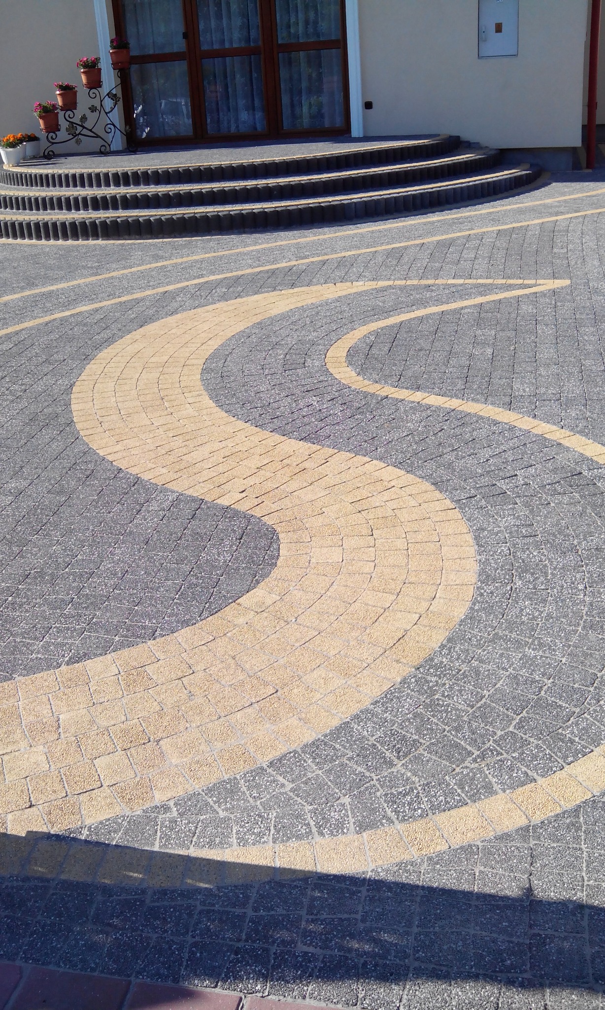 Ułożona kostka brukowa Arko w kolorze szarym i beżowym, tworząca falujący wzór przed wejściem do domu z obrzeżami 6x20 i palisadą Ring, donice z kwiatami na schodach.