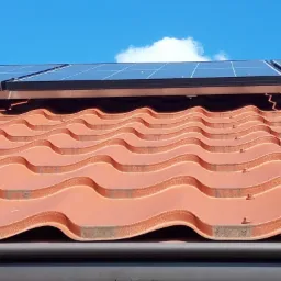 Panele słoneczne zamontowane na dachu z czerwonej dachówki falistej, widok z dołu na tle błękitnego nieba z pojedynczą chmurą.