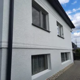 Świeżo wykończona, jasnoszara elewacja domu z widocznymi oknami i ciemnoszarą rynną, ozdobiona poziomymi pasami i kamienną okładziną przy gruncie, na tle błękitnego nieba z chmurami.