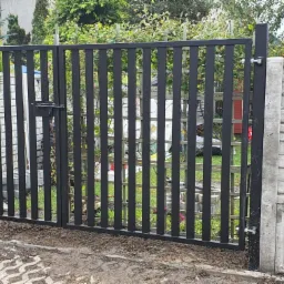 Dwuskrzydłowa brama posesyjna z pionowych profili, czarna, zamontowana między betonowymi słupkami o fakturze cegły, widok na ogród.