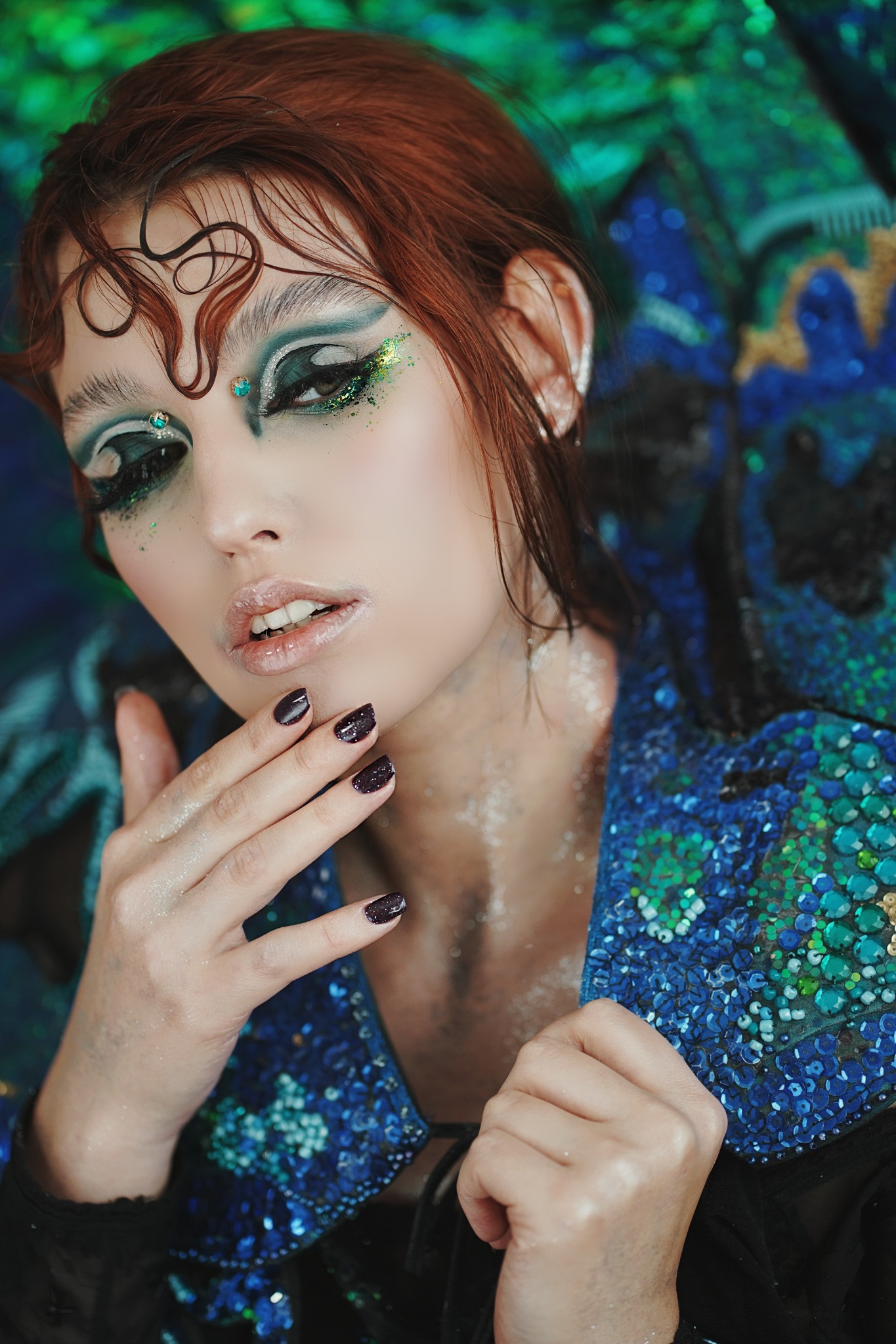 Portret kobiety z artystycznym makijażem oczu w odcieniach zieleni i brokatu, z błyszczącymi ustami i ciemnymi paznokciami, ubrana w cekinową kurtkę.