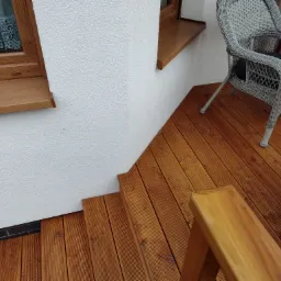 Schody i fragment tarasu z desek o ciepłym, brązowym odcieniu, prowadzące do białej ściany z oknem i wiklinowym krzesłem z szarą poduszką.