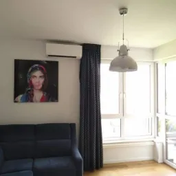 Biały klimatyzator ścienny zamontowany nad obrazem w salonie z granatową sofą i dużym oknem