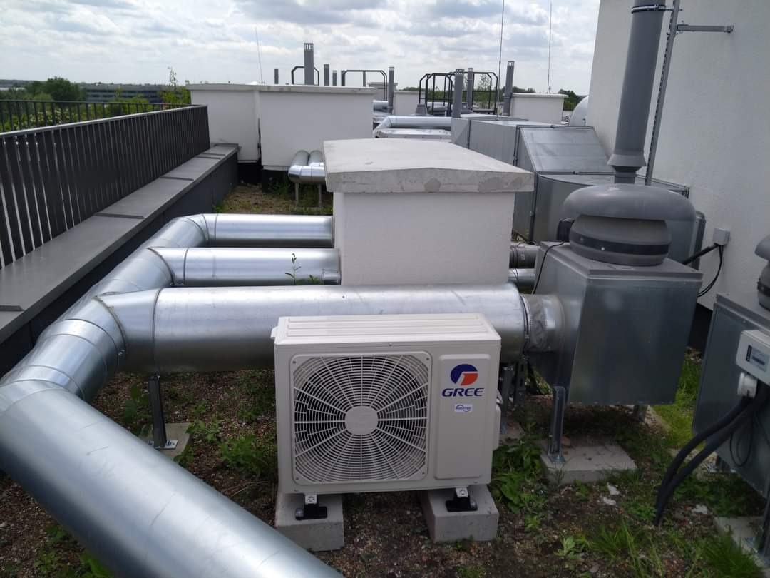 Instalacja klimatyzacyjno-wentylacyjna na dachu budynku komercyjnego, widoczne jednostki zewnętrzne klimatyzacji marki Gree, srebrne rury wentylacyjne i elementy centrali wentylacyjnej.