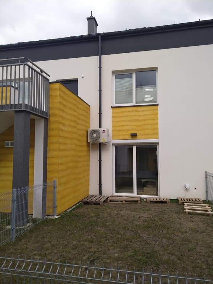 Biała jednostka zewnętrzna klimatyzacji zamontowana na elewacji budynku z żółtymi elementami dekoracyjnymi, widoczna instalacja odprowadzająca skropliny, teren wokół wyłożony drewnianymi paletami.