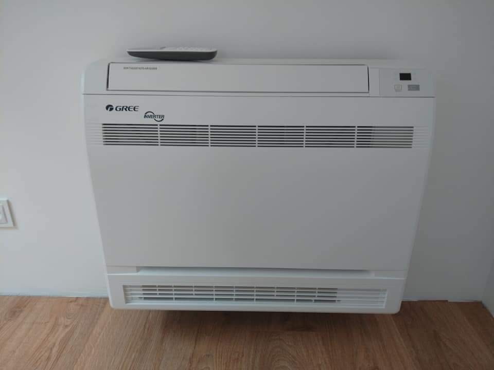 Biały klimatyzator podłogowy marki Gree Inverter z pilotem na górze, zamontowany przy białej ścianie w pomieszczeniu z drewnianą podłogą.