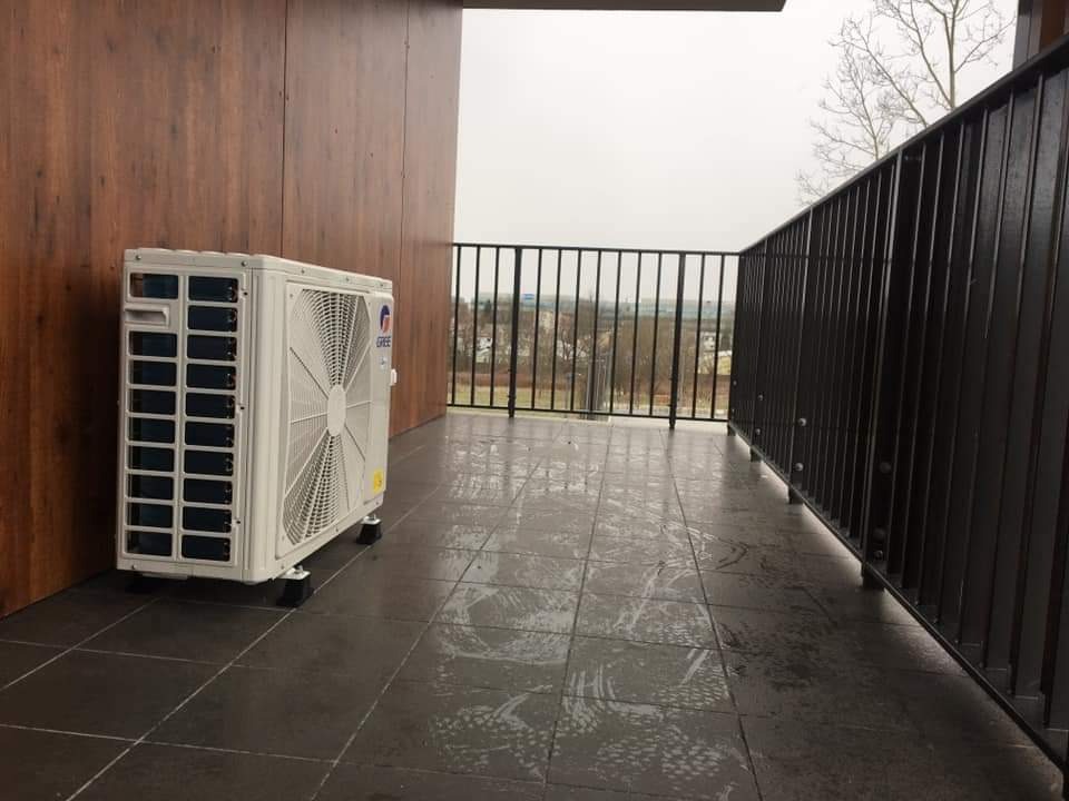 Biała jednostka zewnętrzna klimatyzacji marki Gree na balkonie z ciemną podłogą i balustradą; widok na szary krajobraz za balustradą.