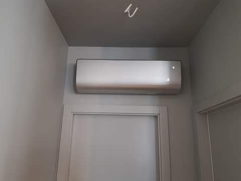 Biały klimatyzator ścienny zamontowany nad drzwiami w pomieszczeniu o szarych ścianach i suficie, widoczne elementy instalacji elektrycznej na suficie.