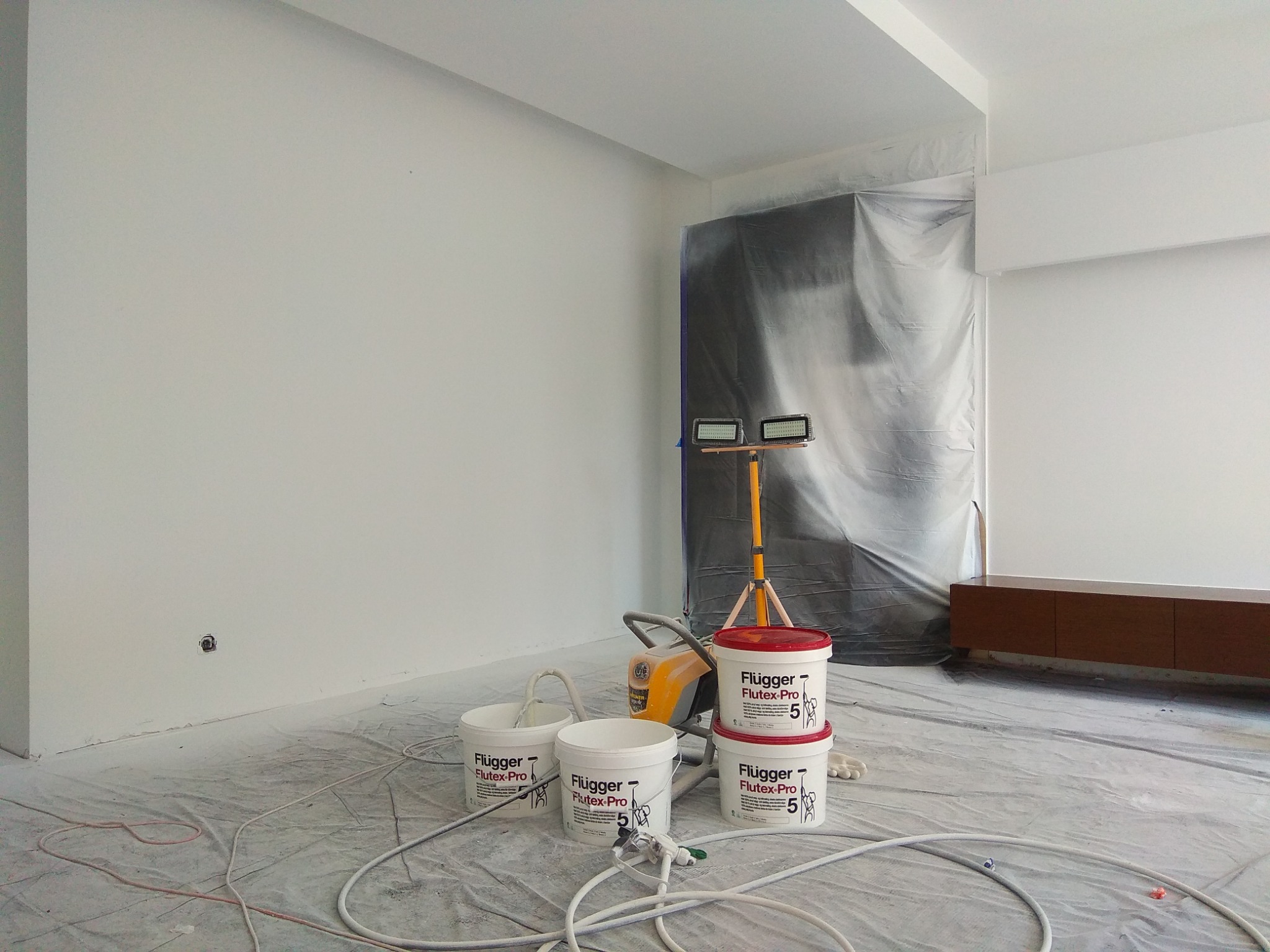Przygotowania do malowania ścian w pomieszczeniu: folia ochronna na podłodze, zakryte meble, reflektor na statywie, agregat malarski i wiadra z farbą Flugger Flutex Pro 5.