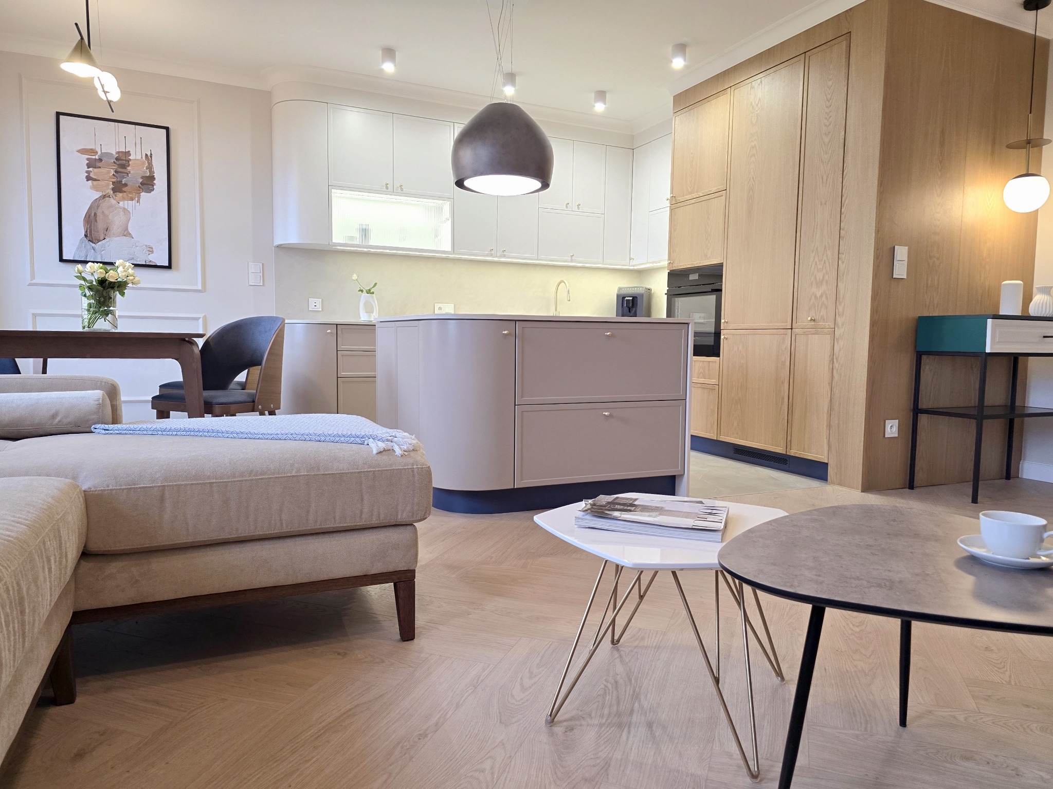 Jasna kuchnia z wyspą i drewnianymi szafkami w nowoczesnym mieszkaniu. Na pierwszym planie stoliki kawowe i fragment sofy. Parkiet w jodełkę.