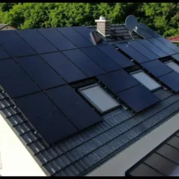 Czarny dach z panelami fotowoltaicznymi i oknami dachowymi, komin z cegły i antena satelitarna, widok z góry na dom jednorodzinny.