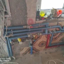 Instalacja hydrauliczna z niebieskimi rurami w trakcie montażu, widoczne zawory, złączki, licznik wody, fragment okna z akcesoriami, ściana z częściowo położonymi płytkami i wiszące przewody...