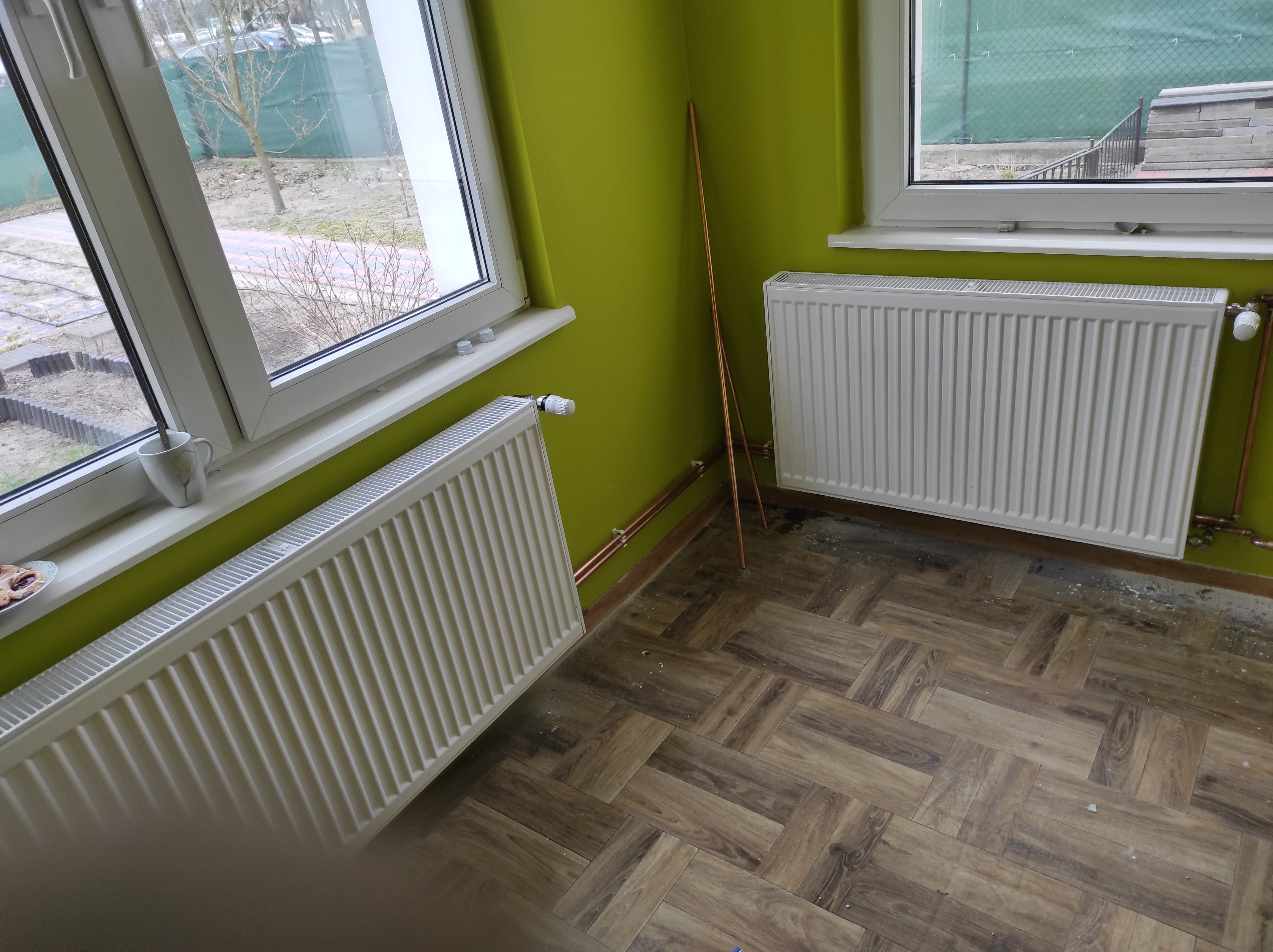 Dwa białe grzejniki zamontowane pod oknami na tle zielonej ściany, z widocznymi miedzianymi rurami instalacji grzewczej i parkietem ułożonym w jodełkę.