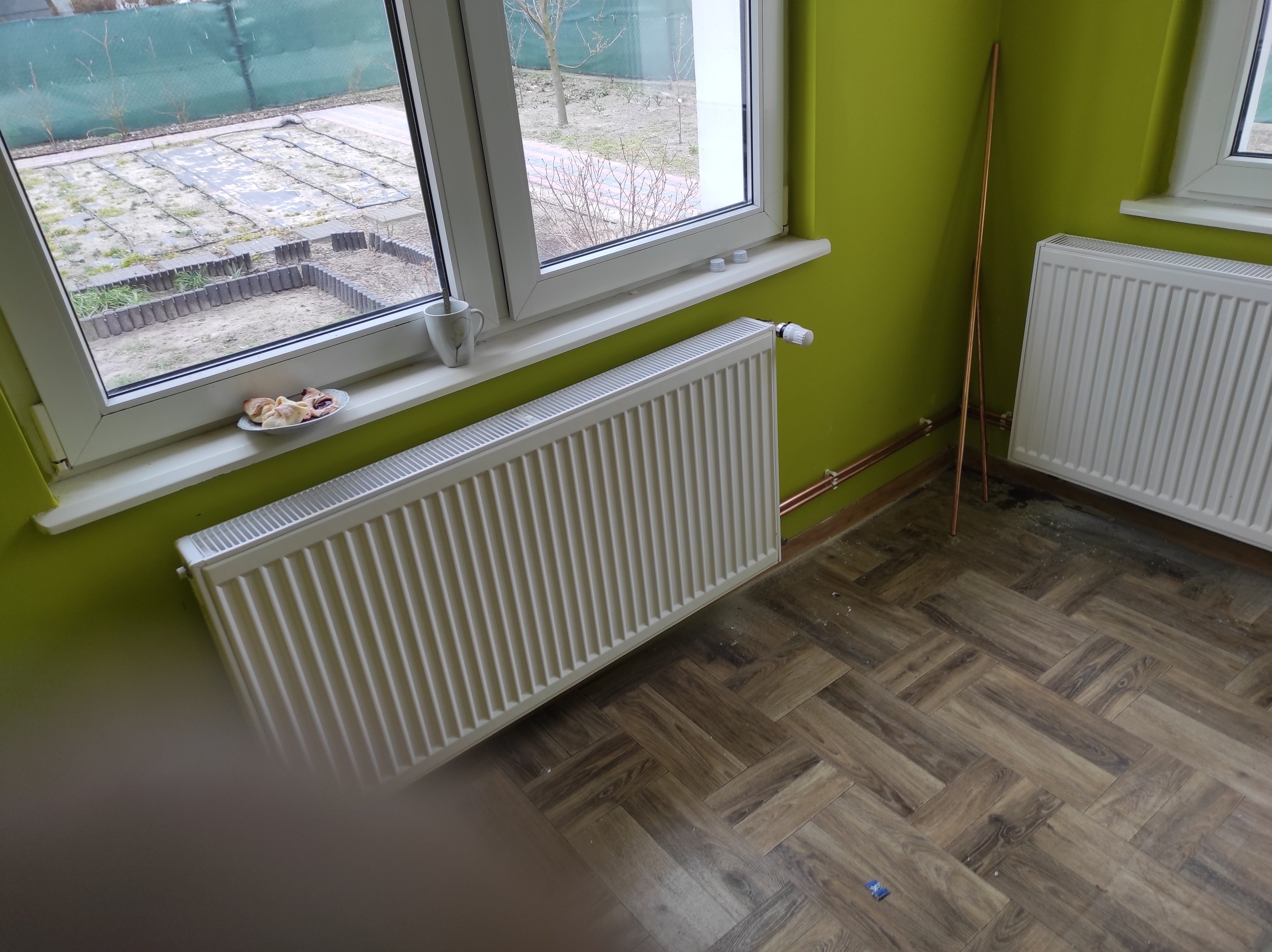 Biały grzejnik panelowy zawieszony pod oknem z parapetem, na którym stoi filiżanka i talerzyk z ciastkami, na tle zielonej ściany i podłogi w jodełkę.