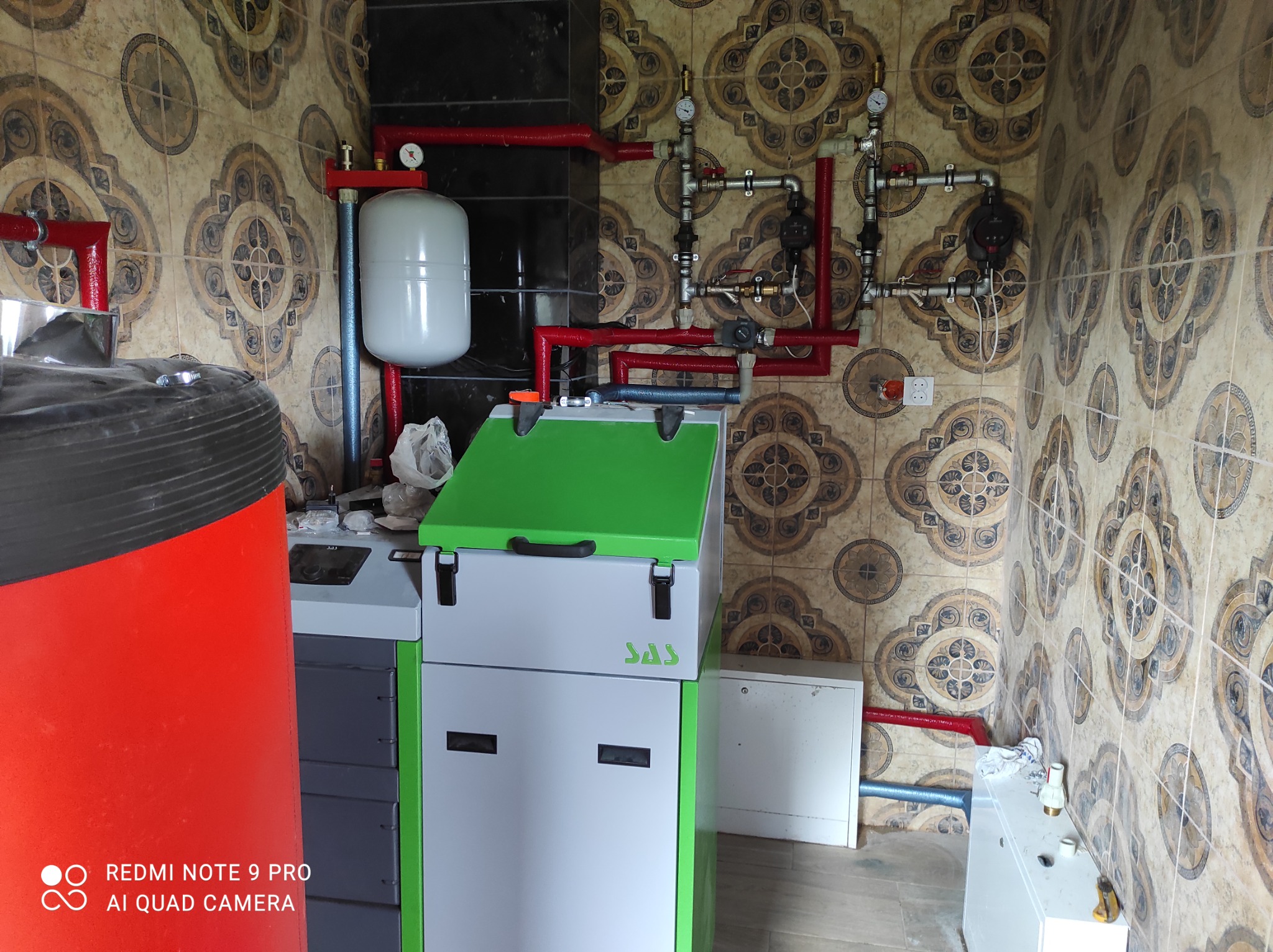 Nowoczesny piec CO marki SAS z zieloną pokrywą, podłączony do instalacji grzewczej z czerwonymi rurami, zainstalowany w pomieszczeniu z dekoracyjnymi płytkami ściennymi.