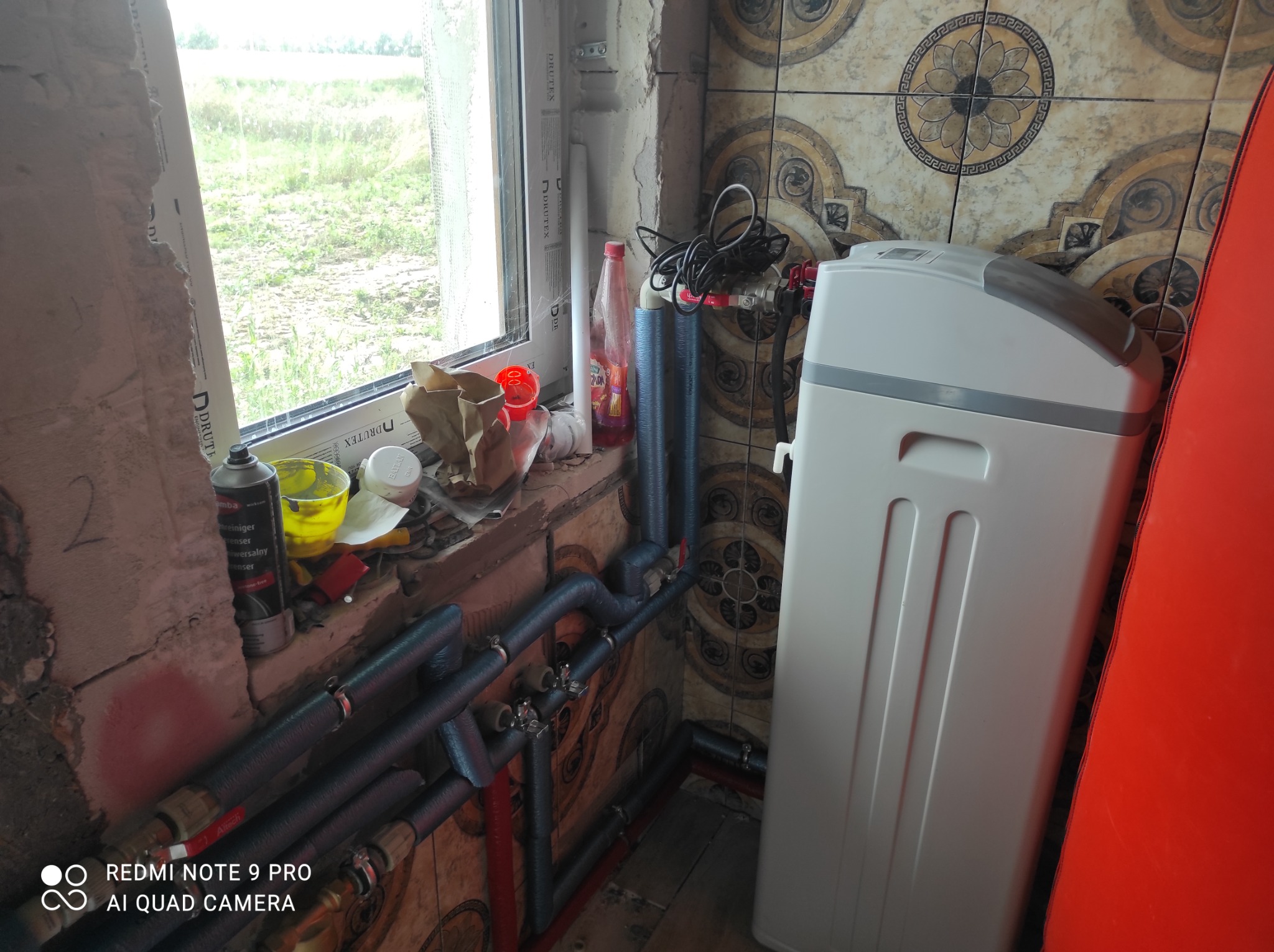 Montaż zmiękczacza wody w pomieszczeniu z częściowo wykończoną ścianą, widoczne rury instalacyjne i okno z widokiem na zewnątrz.