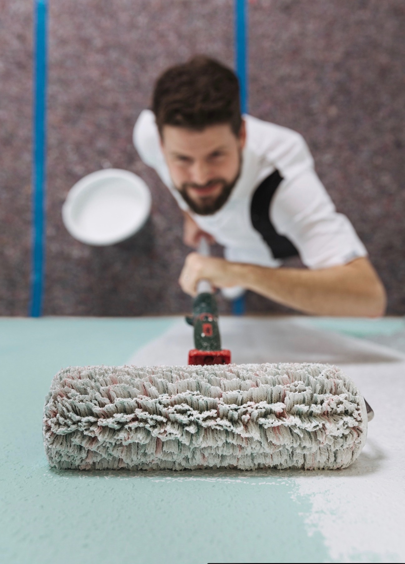 Malowanie ściany wałkiem z bliska, widoczny malarz z uśmiechem i kubełkiem farby w tle.