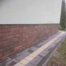 Wykończenie elewacji budynku w Wilczej Woli płytkami imitującymi cegłę, z obrzeżem z kostki brukowej w dwóch kolorach, widok z poziomu gruntu.
