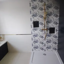 Wykończona łazienka z kabiną prysznicową i złotą armaturą na tle ściany w biało-niebieskie kwiaty, obok ściana z białych płytek z poziomym złotym akcentem.