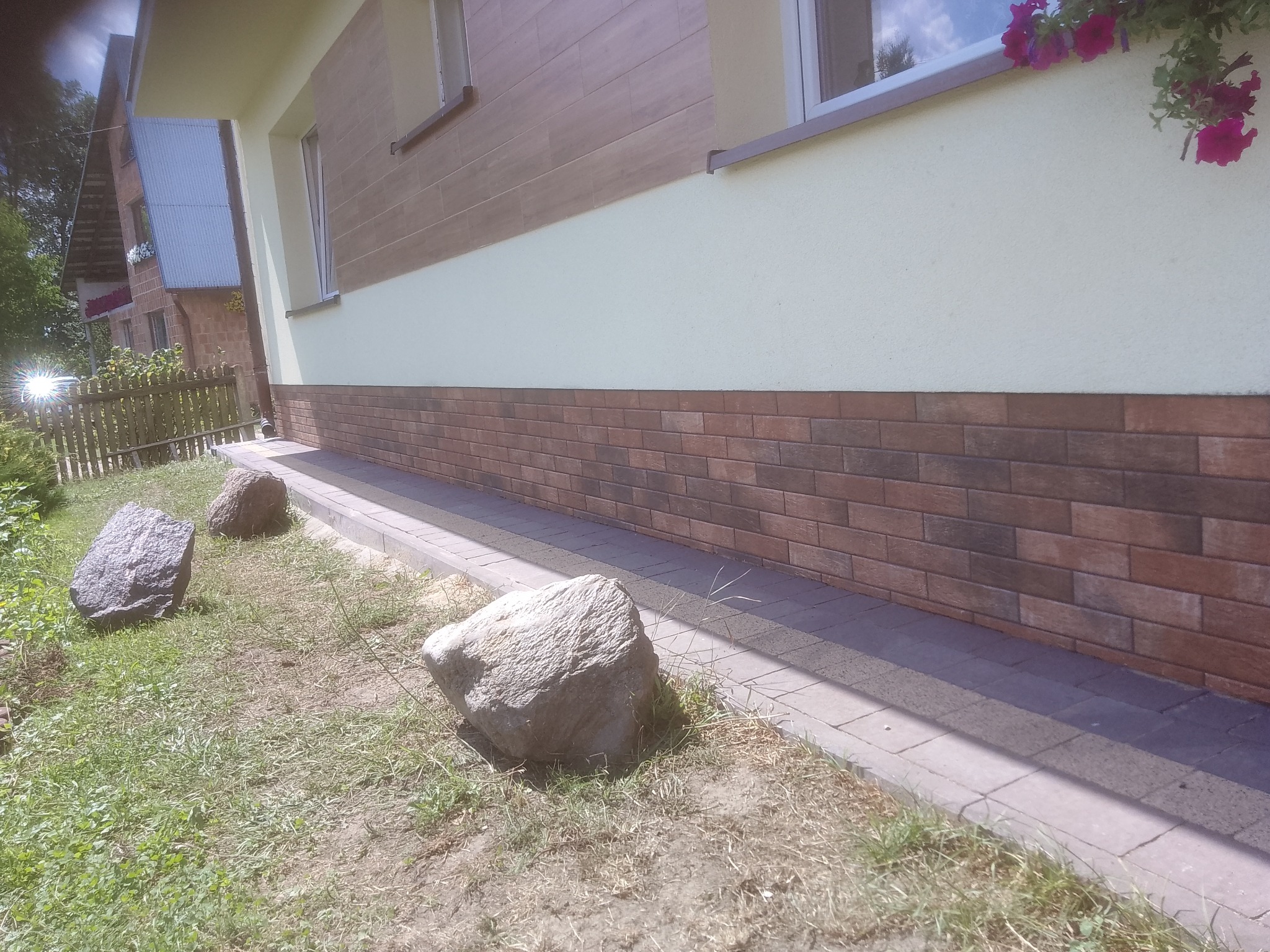 Elewacja domu z brązowymi płytkami imitującymi cegłę na fundamencie, kremowa ściana z oknem i kwiatami, trzy duże kamienie na trawniku.