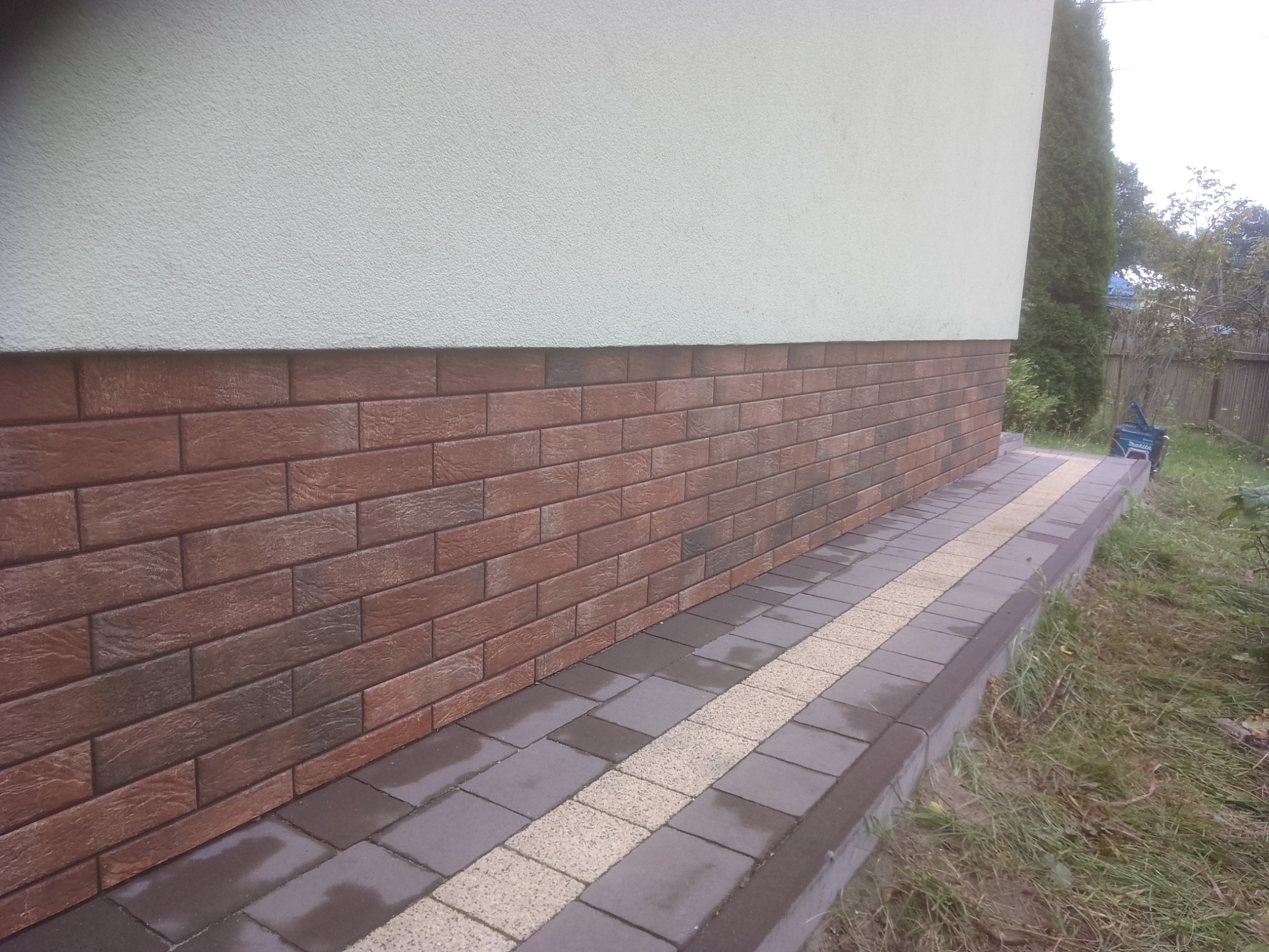 Wykończenie elewacji budynku w Wilczej Woli płytkami imitującymi cegłę, z obrzeżem z kostki brukowej w dwóch kolorach, widok z poziomu gruntu.