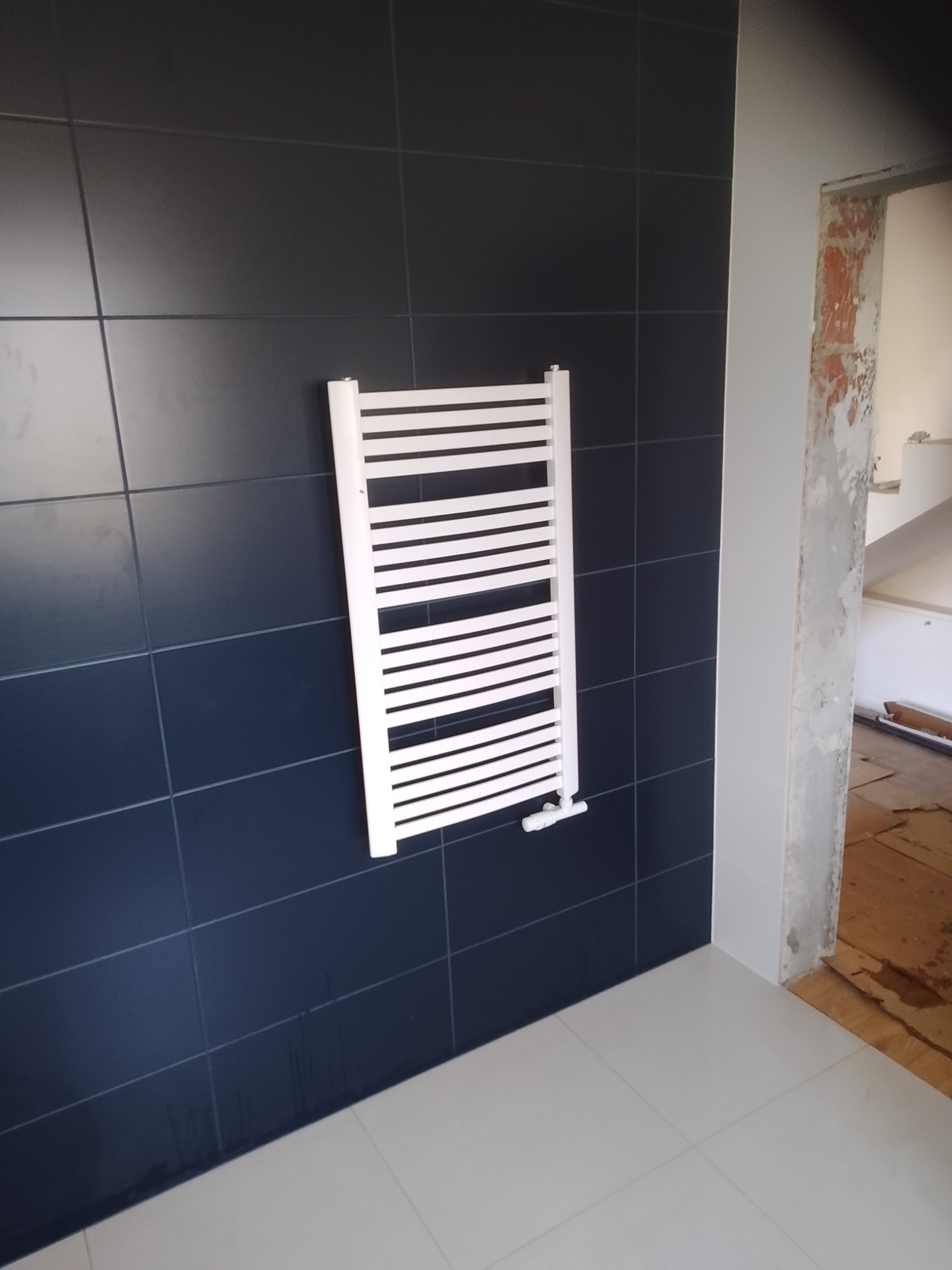 Biały grzejnik drabinkowy zamontowany na ciemnogranatowej ścianie z płytek w łazience w trakcie remontu, widoczne fragmenty nieotynkowanej ściany i podłogi zabezpieczonej kartonem.