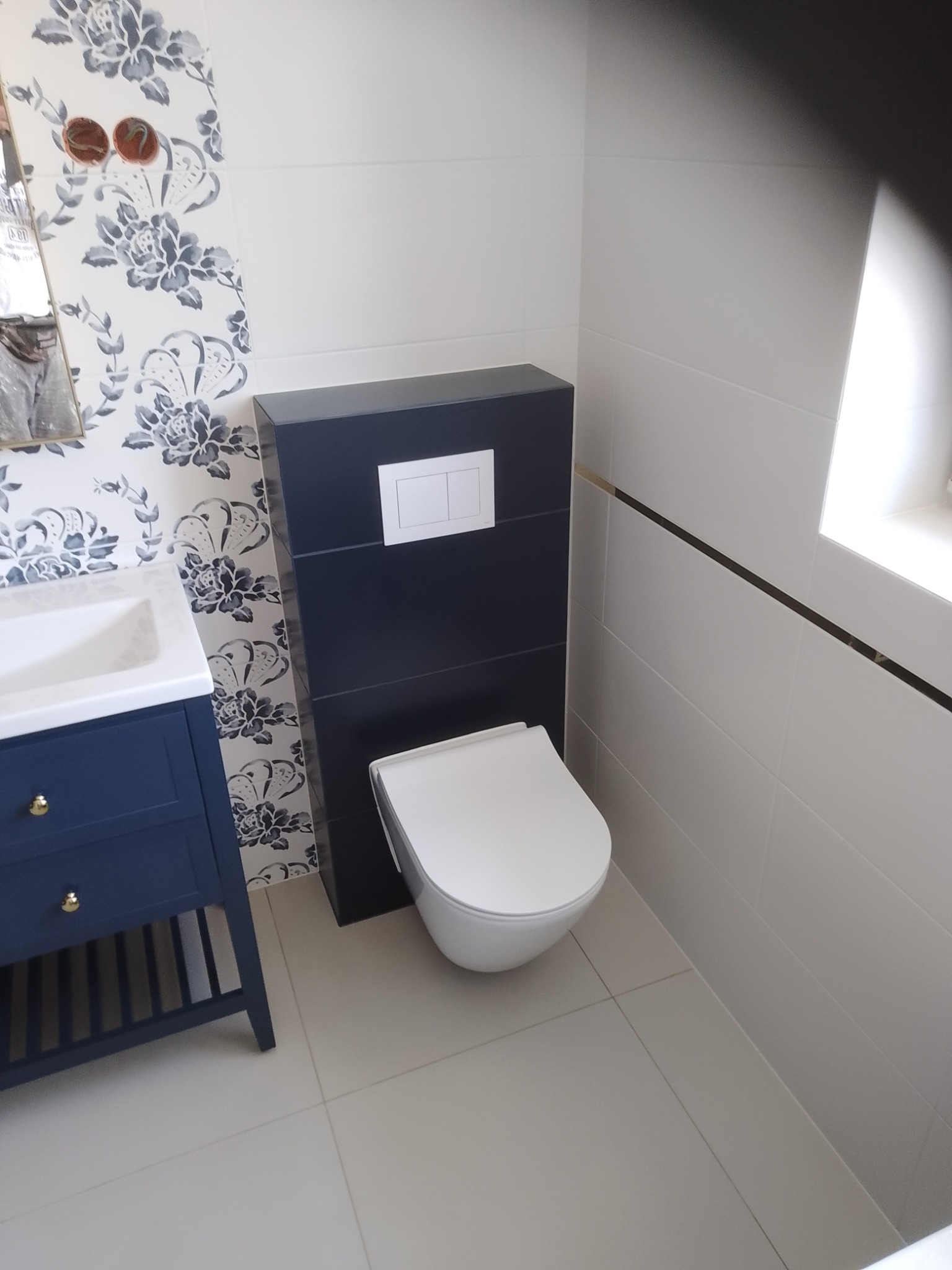 Nowoczesna łazienka z wiszącą toaletą w zabudowie, umywalką z szafką w kolorze granatowym, białymi płytkami z poziomym złotym paskiem i ścianą w kwiatowy wzór, widoczne wystające przewody elektryczne.