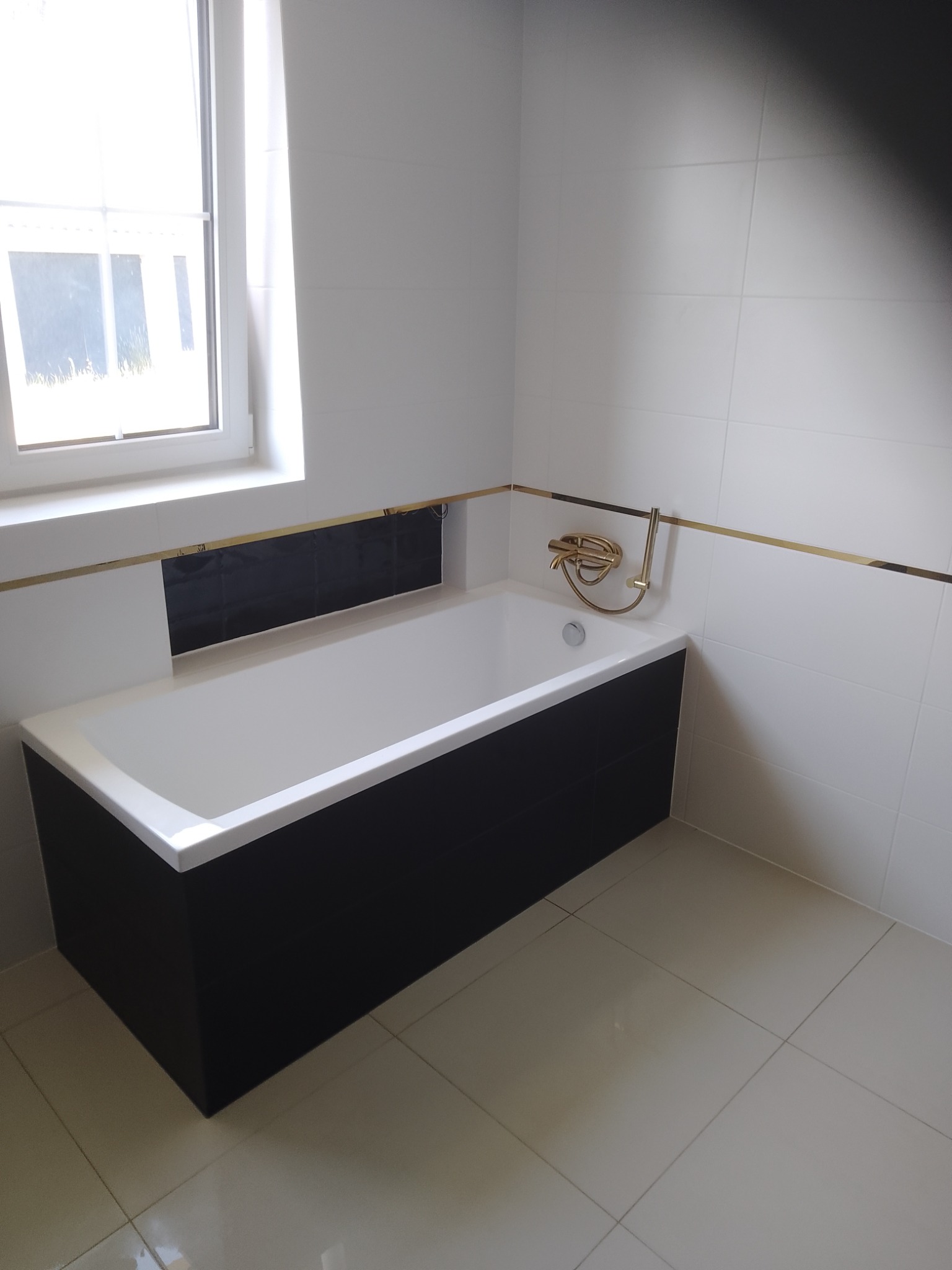 Nowoczesna łazienka z biało-czarną wanną obudowaną ciemnymi panelami, złotą armaturą i listwą dekoracyjną, jasne płytki podłogowe i ścienne, okno z białymi ramami.