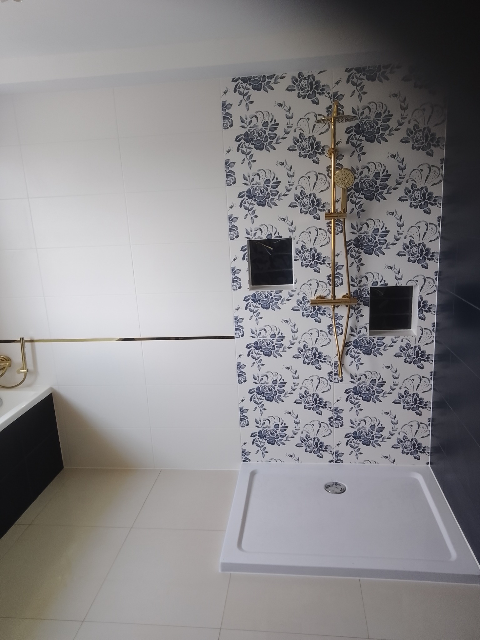 Wykończona łazienka z kabiną prysznicową i złotą armaturą na tle ściany w biało-niebieskie kwiaty, obok ściana z białych płytek z poziomym złotym akcentem.