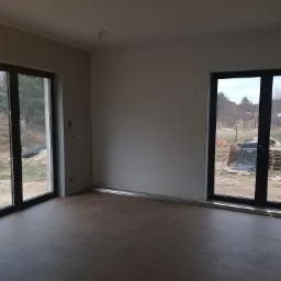 Wnętrze pokoju z ułożonymi panelami podłogowymi w odcieniach brązu, widoczne duże okna balkonowe z czarnymi ramami i widok na zewnątrz z elementami budowy.