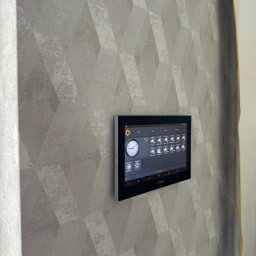 Connect Home Paweł Suchoń - Panel sterowania systemem inteligentnego domu Dahua zamontowany na ścianie z tapetą w geometryczne wzory.