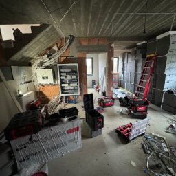 Connect Home Paweł Suchoń - Wnętrze w trakcie prac wykończeniowych, widoczna rozdzielnica elektryczna, kable, narzędzia oraz materiały izolacyjne Swissopor Lambda, surowy betonowy sufit i ceglane ściany.