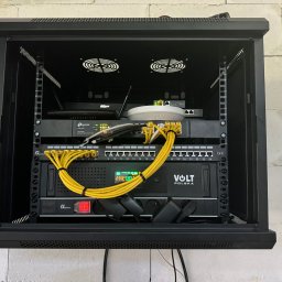 Connect Home Paweł Suchoń - Szafa rack z zainstalowanymi urządzeniami sieciowymi: router, switch, UPS Volt Polska, okablowanie Ethernet w kolorze żółtym, wentylatory chłodzące na górze, zamontowana na ścianie z białej cegły.