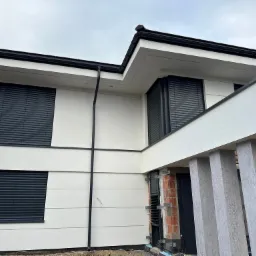 Nowoczesny dom jednorodzinny w trakcie wykończenia z zamontowanymi roletami zewnętrznymi w kolorze antracytowym i elewacją w odcieniach bieli.