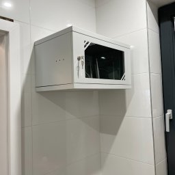 Connect Home Paweł Suchoń - Zamknięta, biała szafa RACK zamontowana na ścianie z białych płytek, widoczne klucze w zamku i częściowo widoczne okablowanie oraz urządzenia wewnątrz szafy, obok widoczna rama okienna.