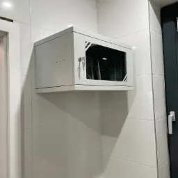 Zamknięta, biała szafa RACK zamontowana na ścianie z białych płytek, widoczne klucze w zamku i częściowo widoczne okablowanie oraz urządzenia wewnątrz szafy, obok widoczna rama okienna.