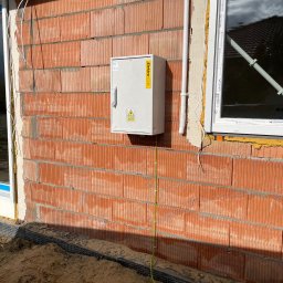 Connect Home Paweł Suchoń - Tymczasowa skrzynia przyłącza budowlanego z uzbrojoną tablicą, zamontowana na ceglanej ścianie budynku w trakcie budowy, widoczne przewody i uziemienie.