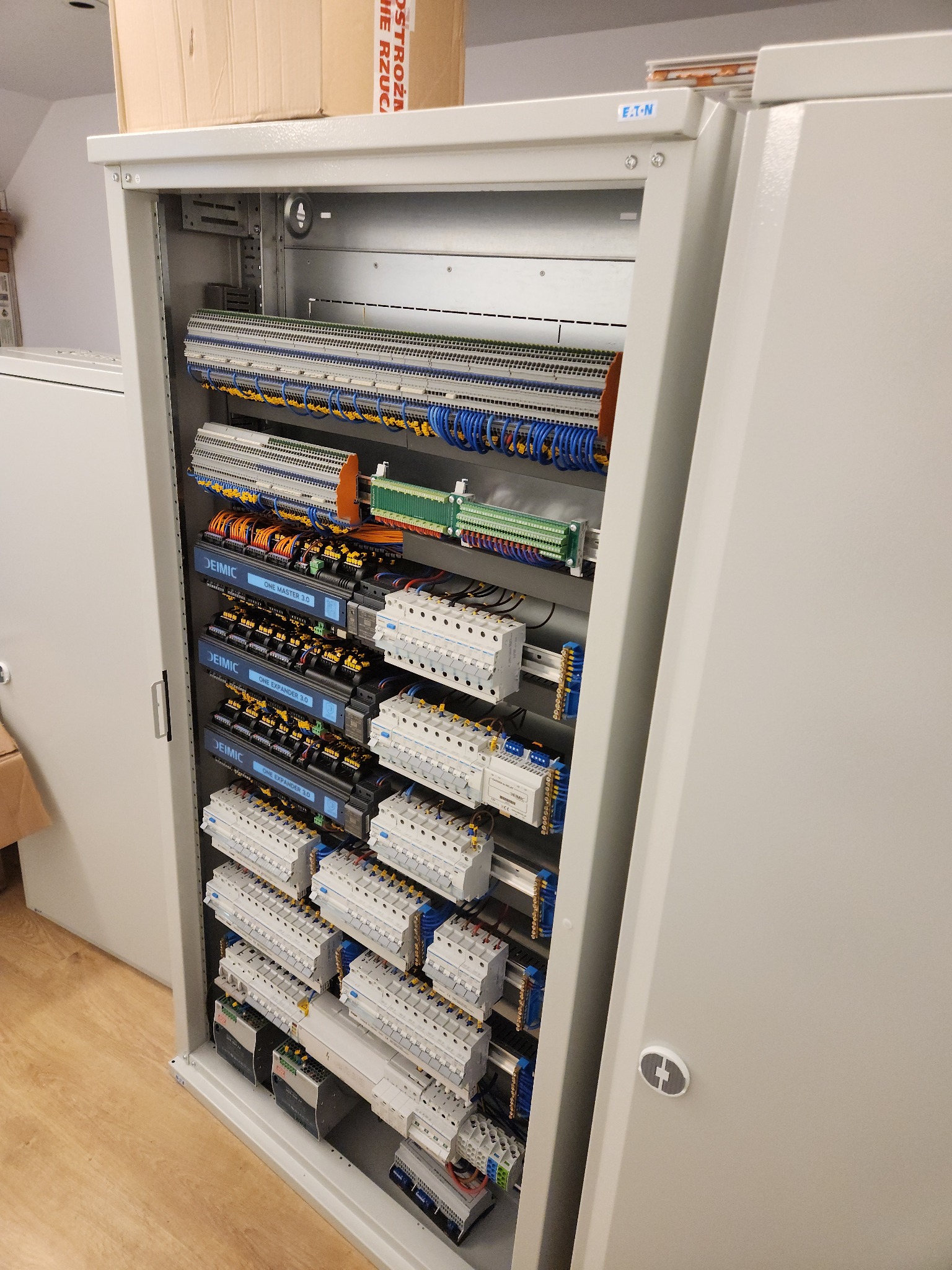 Otwarta szafa sterownicza z widocznymi modułami DEIMIC, licznymi bezpiecznikami, listwami zaciskowymi i okablowaniem w kolorach niebieskim, żółtym i pomarańczowym. Szafa marki EATON.