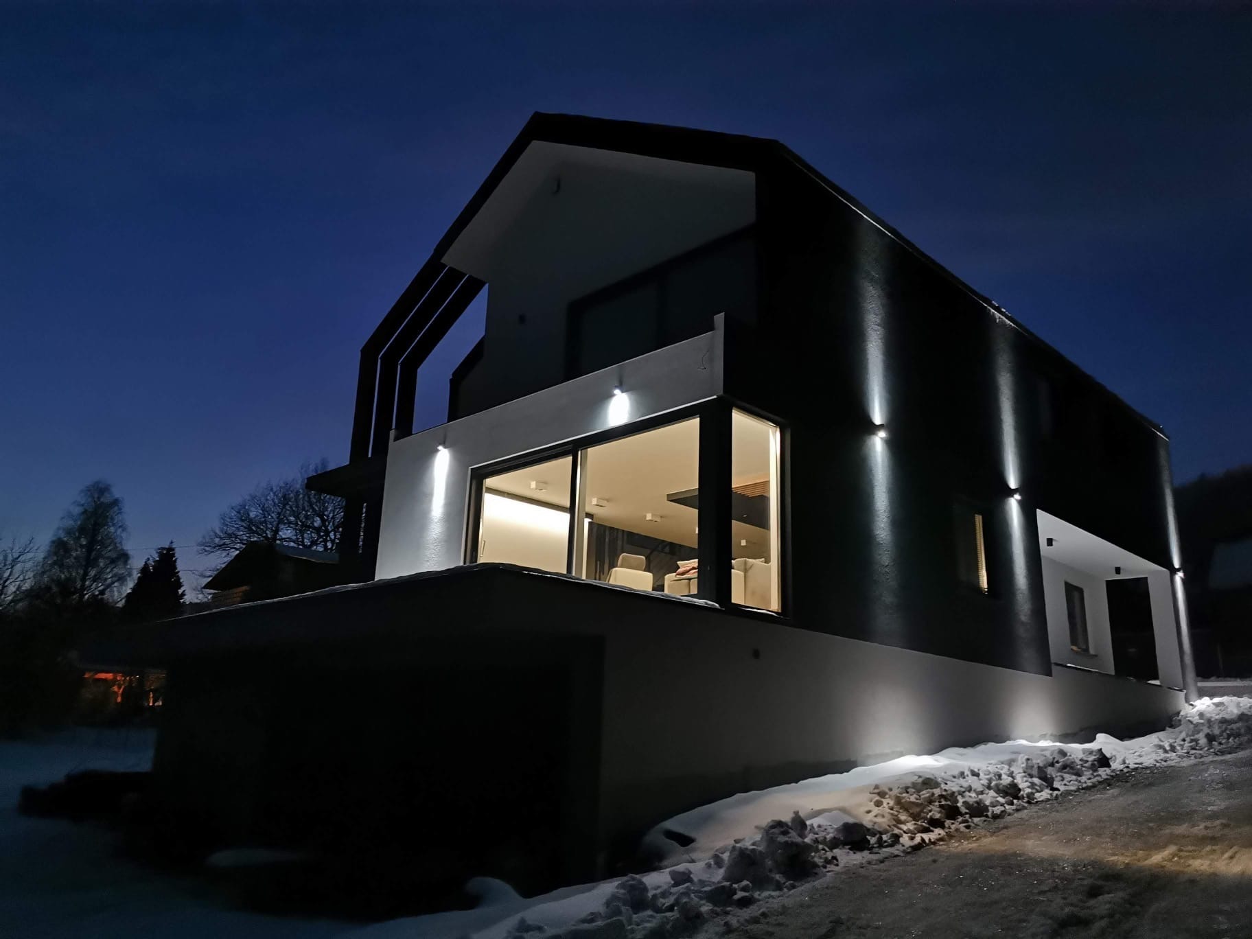 Nowoczesny dom jednorodzinny oświetlony zewnętrznymi lampami punktowymi, widok nocny z zasnieżonym podjazdem i fragmentem ogrodu.
