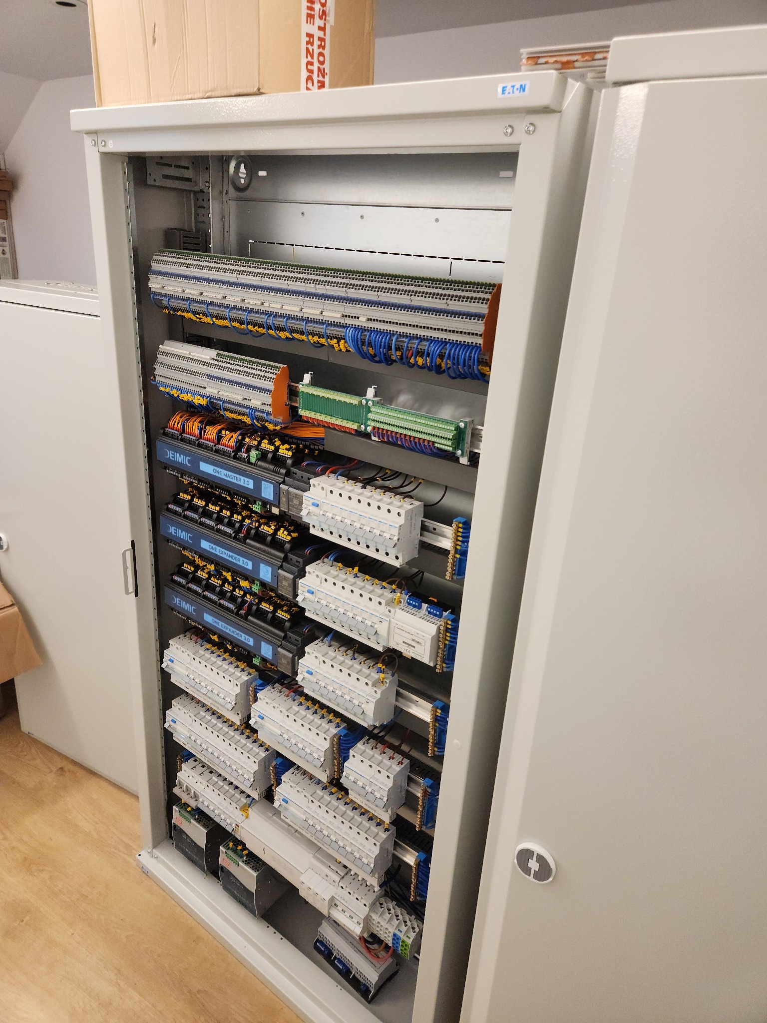 Otwarta szafa elektryczna z widocznymi modułami sterującymi Deimic One Master 3.0 i One Expander 3.0, licznymi bezpiecznikami i rozdzielnicami, okablowaniem w kolorach niebieskim, pomarańczowym...