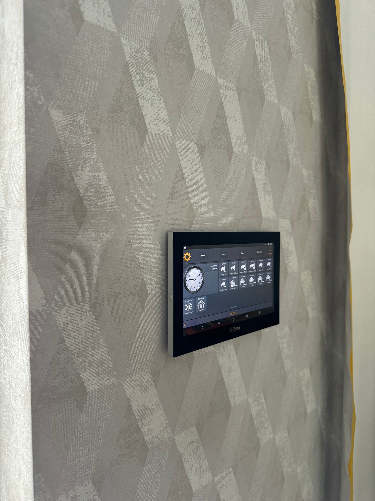 Panel sterowania systemem inteligentnego domu Dahua zamontowany na ścianie z tapetą w geometryczne wzory.