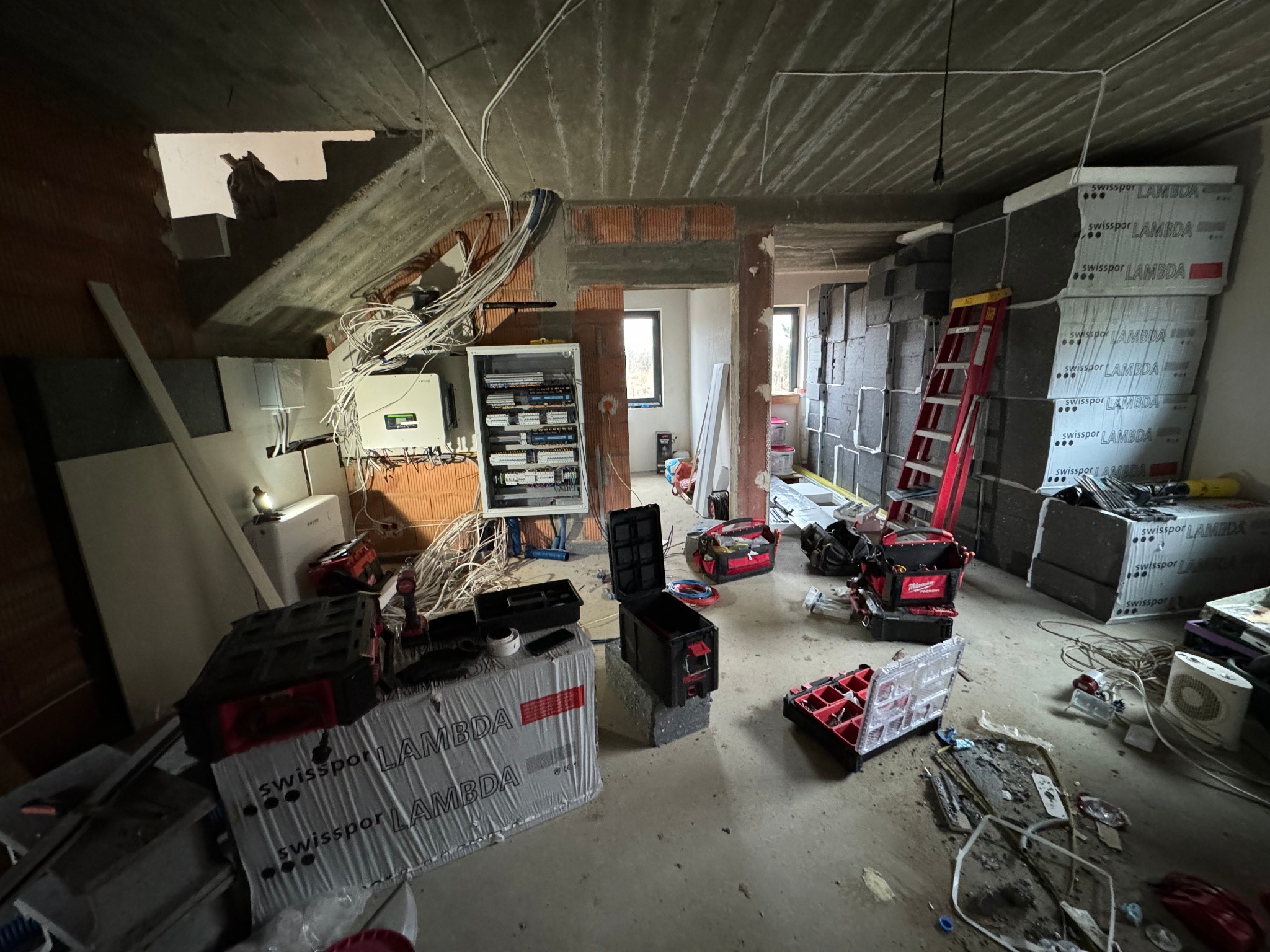 Wnętrze w trakcie prac wykończeniowych, widoczna rozdzielnica elektryczna, kable, narzędzia oraz materiały izolacyjne Swissopor Lambda, surowy betonowy sufit i ceglane ściany.