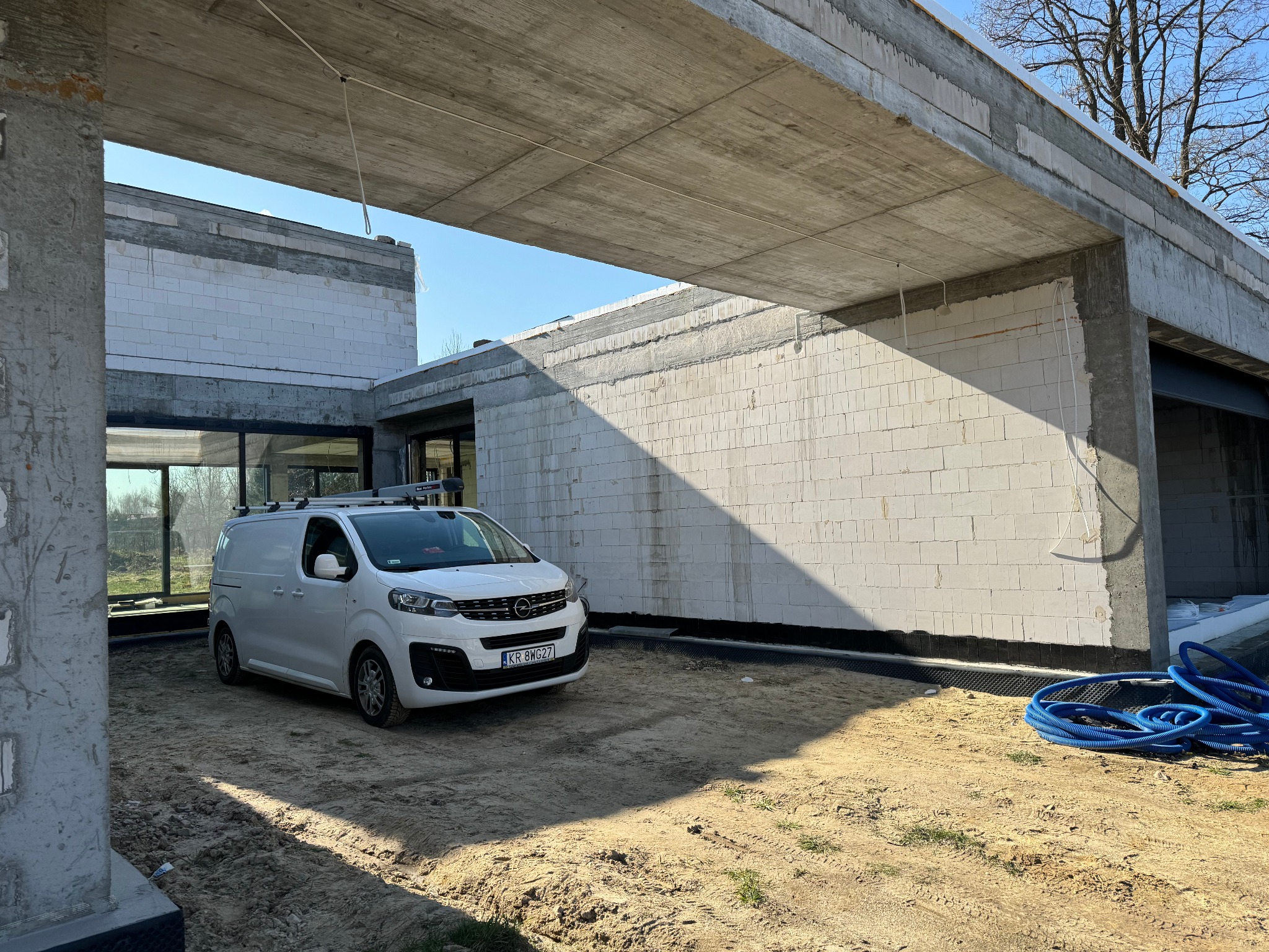 Biały samochód dostawczy z bagażnikiem dachowym na tle surowego stanu budynku z białego betonu komórkowego i betonowej konstrukcji stropu.