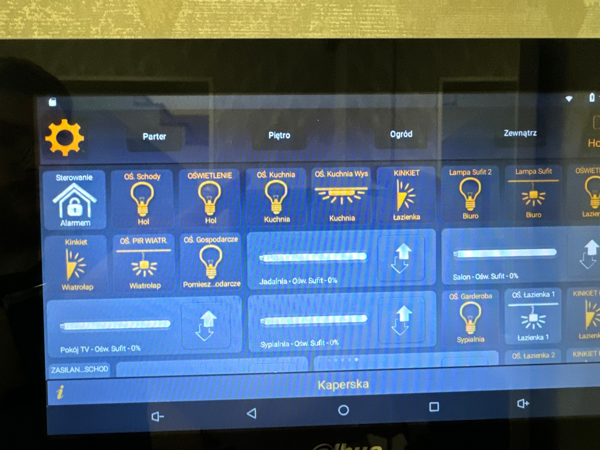 Ekran tabletu z interfejsem systemu zarządzania oświetleniem w domu, widoczne ikony i nazwy pomieszczeń: parter, piętro, ogród, hol, kuchnia, łazienka, biuro, sypialnia, wiatrołap, jadalnia, salon,...