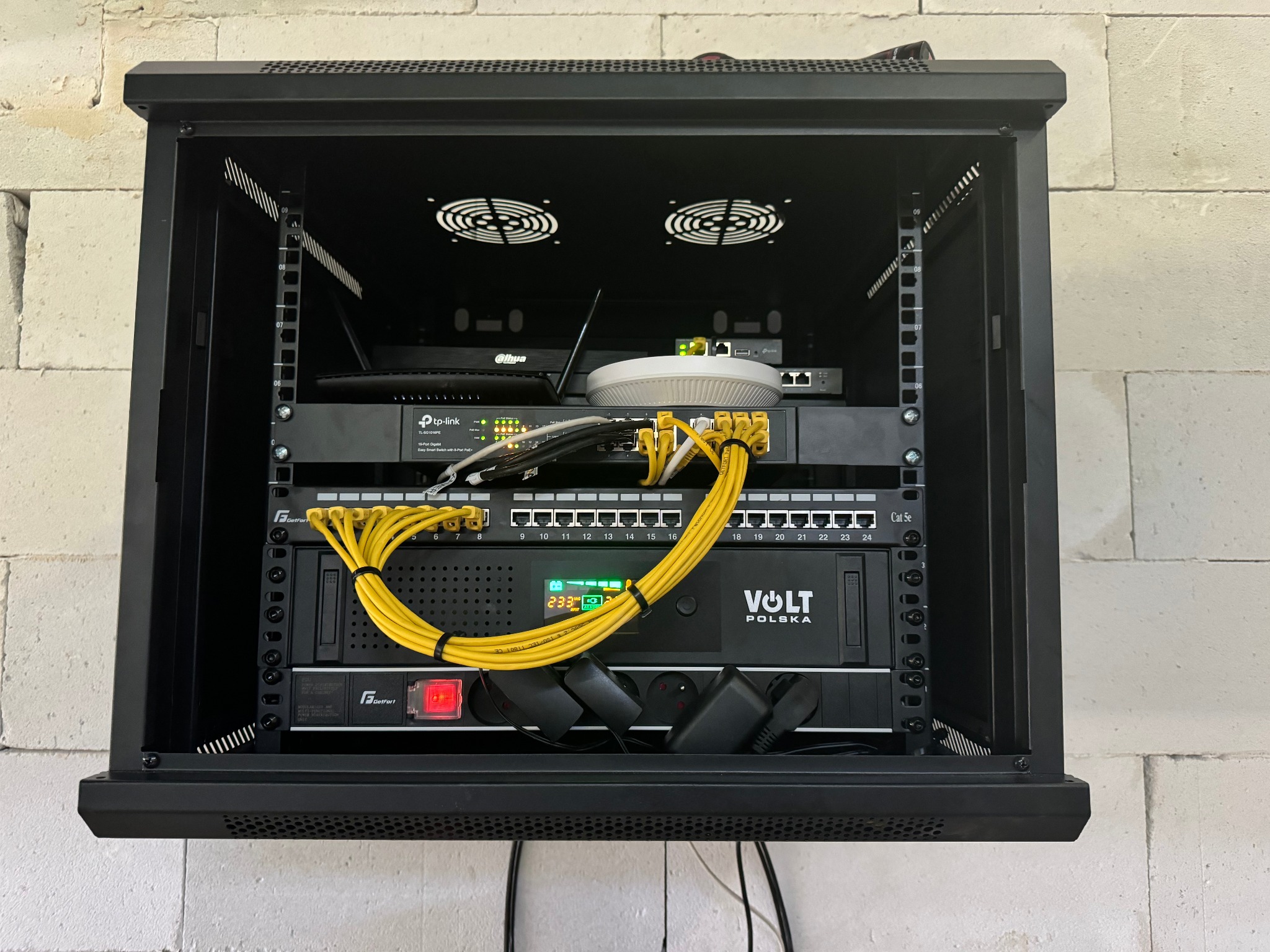 Szafa rack z zainstalowanymi urządzeniami sieciowymi: router, switch, UPS Volt Polska, okablowanie Ethernet w kolorze żółtym, wentylatory chłodzące na górze, zamontowana na ścianie z białej cegły.