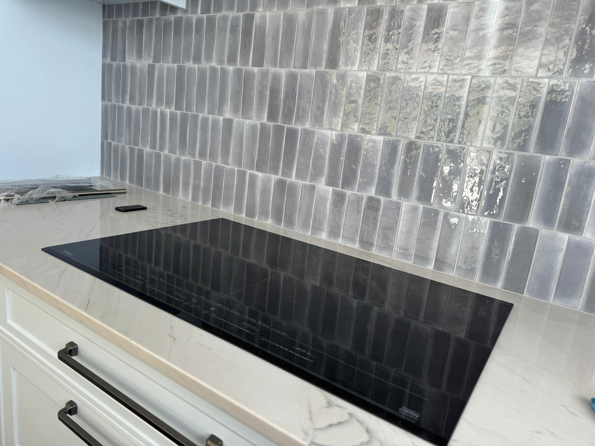 Nowoczesna płyta indukcyjna wkomponowana w marmurowy blat kuchenny, z tłem z płytek w odcieniach szarości ułożonych pionowo.